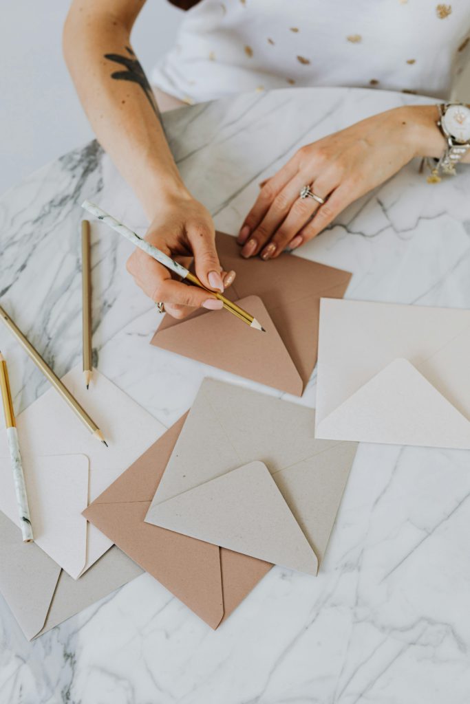 Flat lay of hands writing on envelopes with pencils on marble surface.